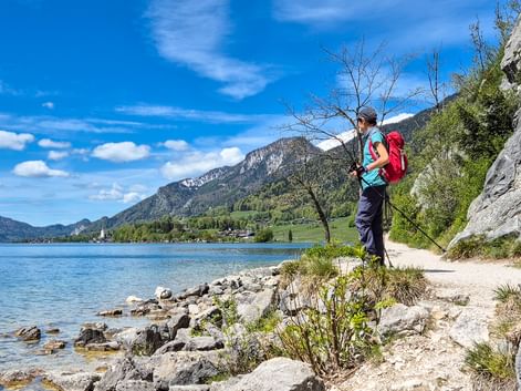 Female hiker with red backpack standing on rocky shore of Wolfgangsee lake in Salzkammergut, Austria, with mountains and blue sky in background.
