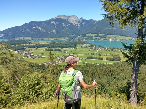 Wanderin mit grünem Rucksack und Wanderstöcken blickt auf den Wolfgangsee und Berge im Salzkammergut. Grüne Täler und Dörfer sind unten sichtbar.