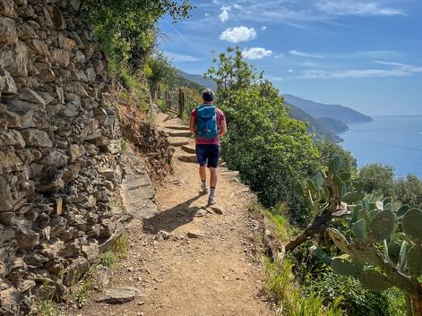 Hiker with blue backpack walking on stone path along Ligurian coast. Stone wall on left, Mediterranean Sea and green hillside on right.