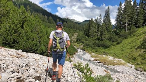 Wanderer mit Rucksack und Stöcken auf felsigem Weg entlang eines Bergbachs am Lechweg, umgeben von Wald und Alpengipfeln.