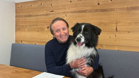Mann namens Andi sitzt mit schwarz-weißem Border Collie namens Finn an Holztisch. Holzverkleidete Wand im Hintergrund, Dokument auf Tisch.