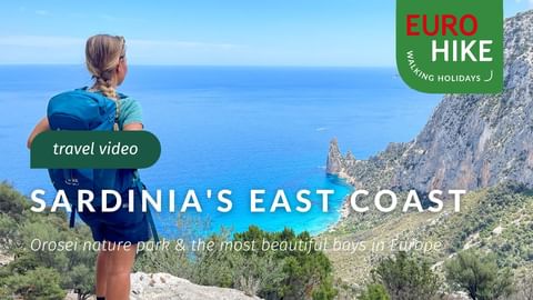 Female hiker with backpack viewing turquoise Mediterranean Sea and rocky coastline in Sardinia. Steep cliffs and green vegetation visible.