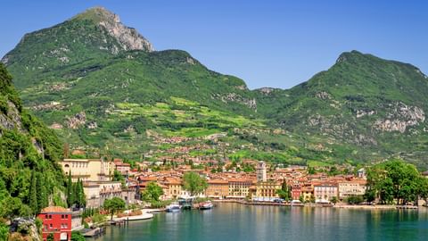 Panoramablick auf Riva del Garda mit bunten Gebäuden entlang des Gardasee-Ufers, umgeben von grünen Bergen unter blauem Himmel.