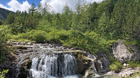Wasserfall, der über felsige Kanten in ein türkisfarbenes Becken am Lechweg fällt, umgeben von üppigem grünem Wald und Bergen.