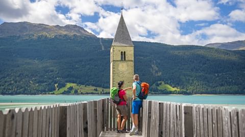 Wanderer blicken auf den türkisen Reschensee mit seinem versunkenen Kirchturm
