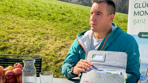 Mann in türkisfarbener Jacke präsentiert graue Reisetasche an Tisch im Freien mit Wasserkaraffe und Äpfeln, grüne Wiese und Wald im Hintergrund.