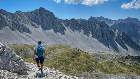 Hiker with backpack walking on rocky mountain path in Karwendel with dramatic jagged peaks and scree slopes under blue sky with clouds.