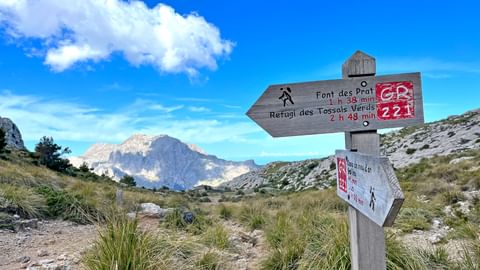 Wooden signpost on GR221 trail showing directions to Font des Prat and Refugi des Tossals Verds, with rocky mountain landscape under blue sky.