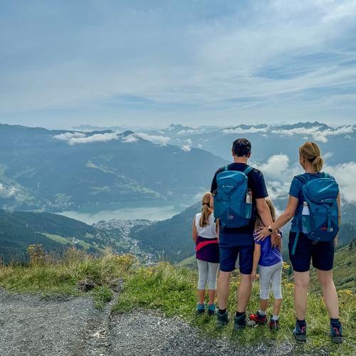 Family of four with backpacks viewing alpine panorama in Pinzgau. Mountain lake and peaks visible below through clouds.