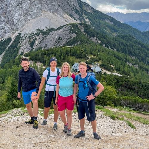 Vier Wanderer mit Rucksäcken auf dem Vršič-Pass-Gipfel mit bewaldeten Bergen und felsigen Gipfeln der Julischen Alpen im Hintergrund.