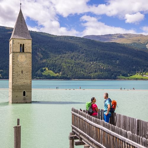 Two hikers on a footbridge in front of the church tower of Altgraun in Lake Reschen