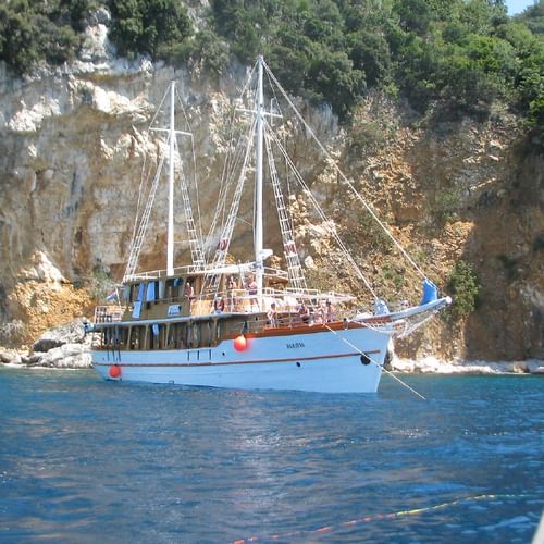 White sailing yacht MS Dalmatino anchored in blue waters near limestone cliffs covered with green vegetation in a Croatian bay.