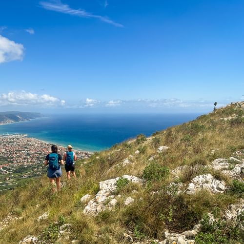 Two hikers walking uphill on Monte Croce with coastal views of Piedmont-Liguria. A summit cross is visible on the rocky peak under blue sky.
