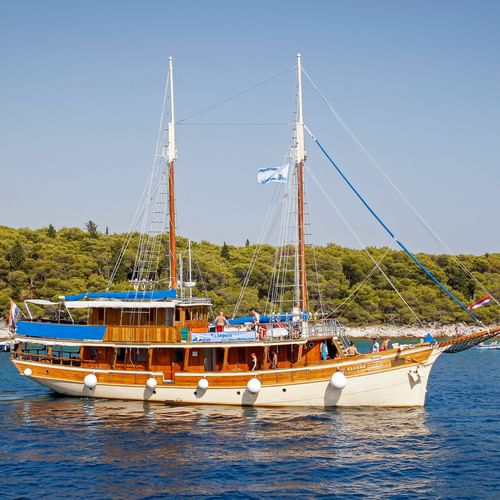 Wooden sailing ship MS Sagena with two masts and blue canopy anchored in calm blue water. Green forested coastline visible in background.