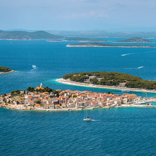 Aerial view of Primošten peninsula with terracotta roofs, church tower, and turquoise Adriatic Sea. Islands and mountains visible in background.