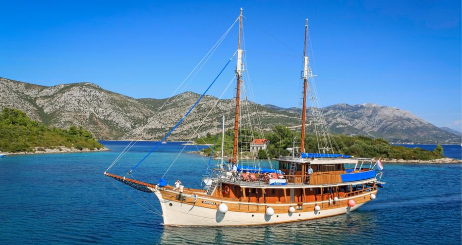 Traditional wooden sailing ship MS Sagena with two masts in turquoise waters of Kvarner Bay, Croatia, with mountains and coastline in background.