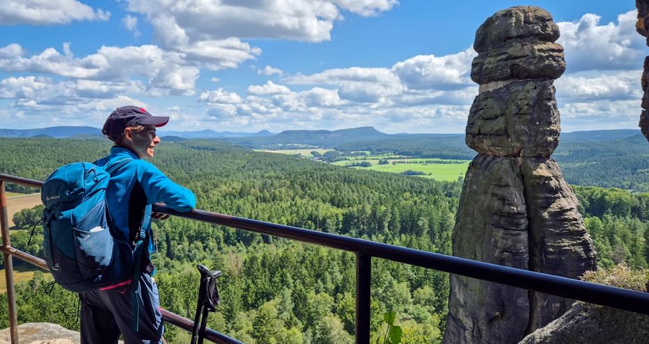 Wanderer mit blauem Rucksack am Pfaffenstein Aussichtspunkt mit Blick auf bewaldete Täler und Felder. Markante Felsformation neben dem Geländer.