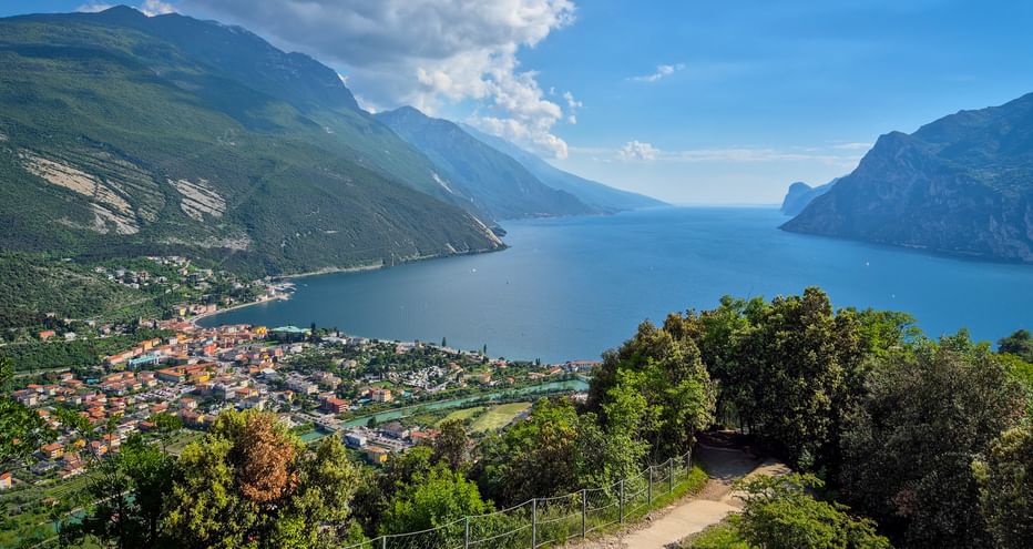 Hiking trail on Monte Brione overlooking Lake Garda, Torbole, and surrounding mountains under blue sky with white clouds.
