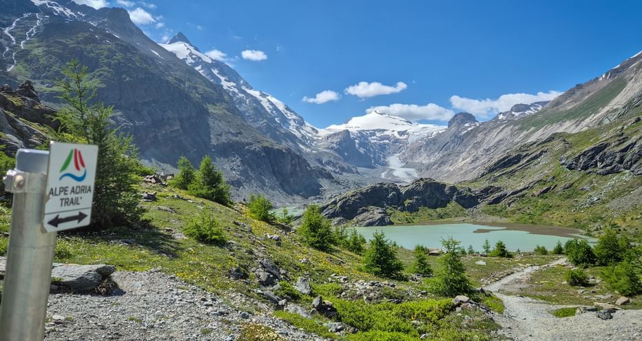 Alpe Adria Trail Wegweiser beim Margaritzensee mit Großglockner Gletscher und schneebedeckten Gipfeln unter blauem Himmel.