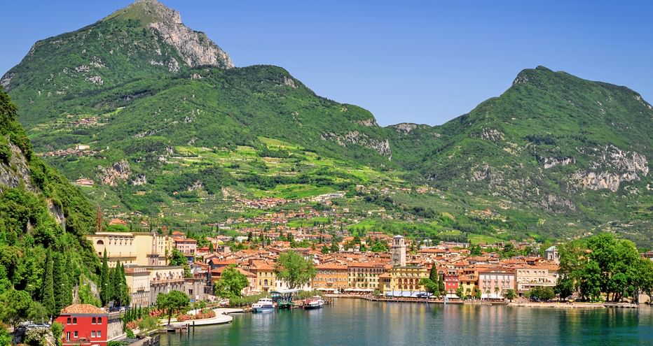 Panoramic view of Riva del Garda with historic buildings along Lake Garda's shoreline, surrounded by green mountains under clear blue sky.