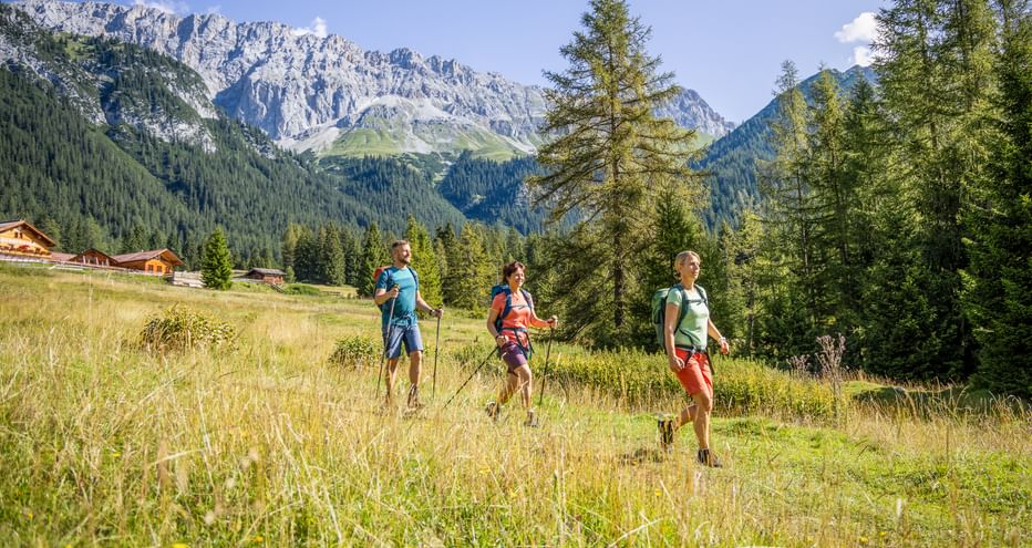Drei Wanderer mit Rucksäcken gehen durch Almwiese im Wettersteingebirge. Felsige Gipfel und traditionelle Holzgebäude sichtbar.