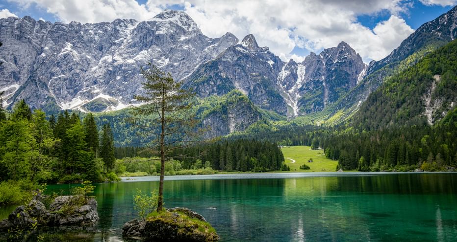 Emerald green lake Laghi di Fusine with small rocky island and lone tree. Snow-capped mountain peaks rise behind dense forests under cloudy sky.