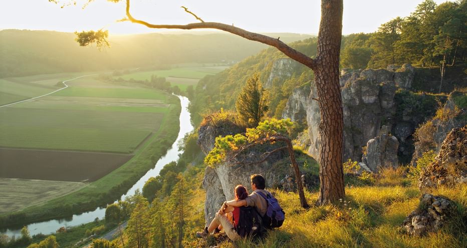 Two hikers sitting on a rocky cliff in Altmühltal, overlooking a winding river through green valley at sunset with pine tree in foreground.