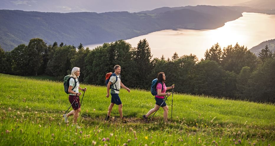 Three hikers with backpacks and walking poles crossing a green meadow in Döbriach, with mountains and a lake visible in the background.
