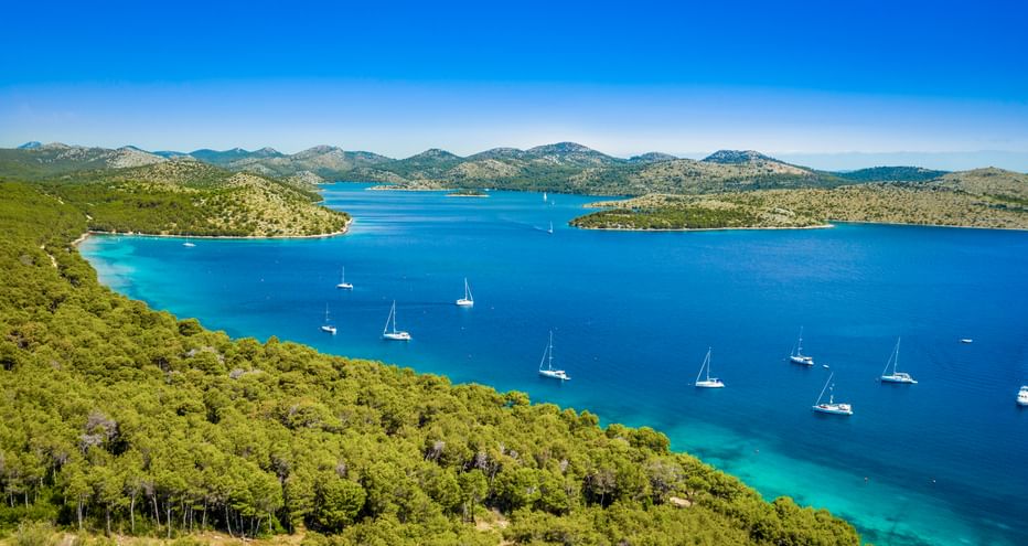 Aerial view of Dugi Otok bay in North Dalmatia with turquoise water, white sailboats, green forested coastline, and mountains under blue sky.