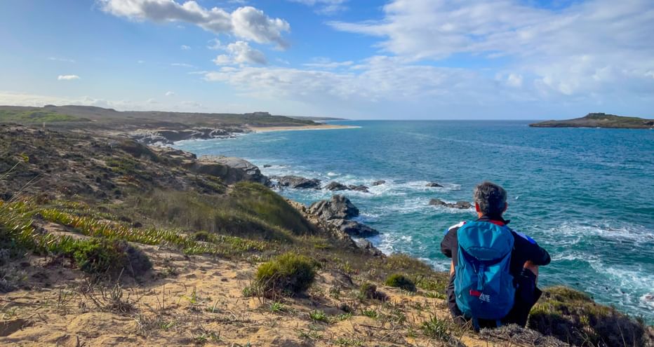 Wanderer mit blauem Rucksack sitzt auf Küstenklippe mit Blick auf türkisfarbenen Atlantik bei Porto Covo, Praia do Sissal, Rota Vicentina, Portugal.