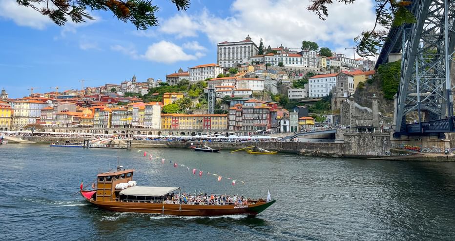 Traditionelles Holzboot mit Passagieren auf dem Douro in Porto, mit bunten Häusern am Hang und der Dom Luís I Brücke.
