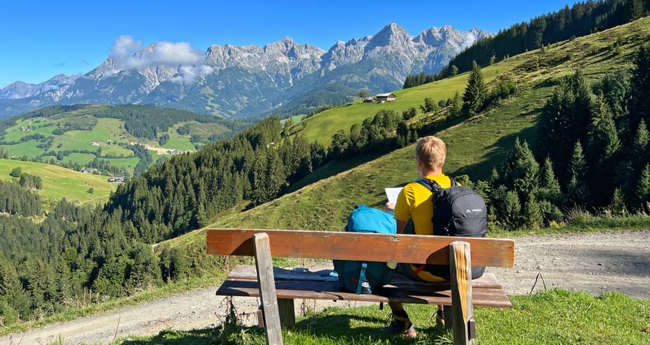 Two hikers with backpacks sit on a wooden bench overlooking green alpine meadows and the rocky Kitzbüheler Alps under a blue sky.