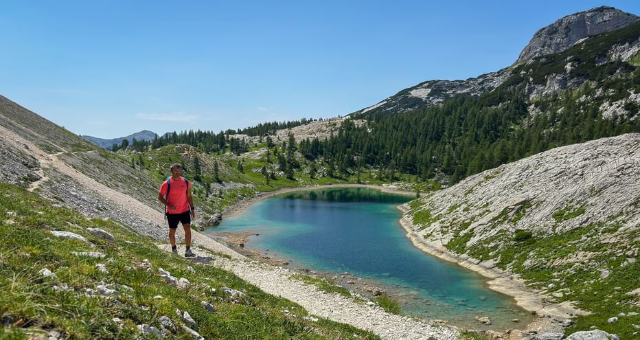 Hiker in red jacket overlooking turquoise alpine lake in Julian Alps valley, surrounded by rocky slopes, green vegetation and mountain peaks.