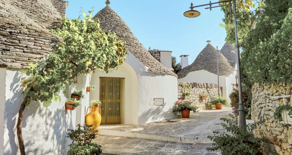 Sonnige Straße in Alberobello mit weiß getünchten Trulli-Häusern mit konischen Steindächern, Blumentöpfen und einem kleinen Baum.