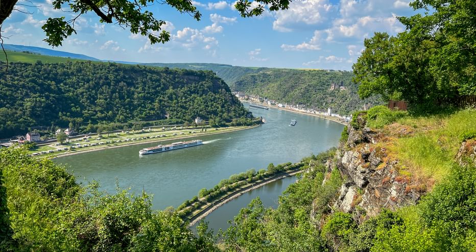 Panoramic view of Rhine River winding through green valley with cruise ship, forested hills, and town along riverbank, seen from Loreley Rock.