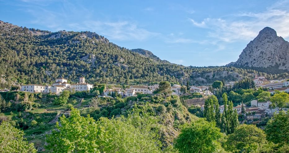 Weiße Dorfhäuser der Alpujarras zwischen grünen Hügeln und Bergen unter blauem Himmel, mit markanter Felsspitze im Hintergrund.