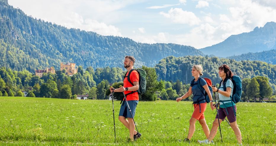 Drei Wanderer mit Rucksäcken gehen über eine grüne Wiese bei Hohenschwangau. Bewaldete Berge und Schloss Hohenschwangau im Hintergrund sichtbar.