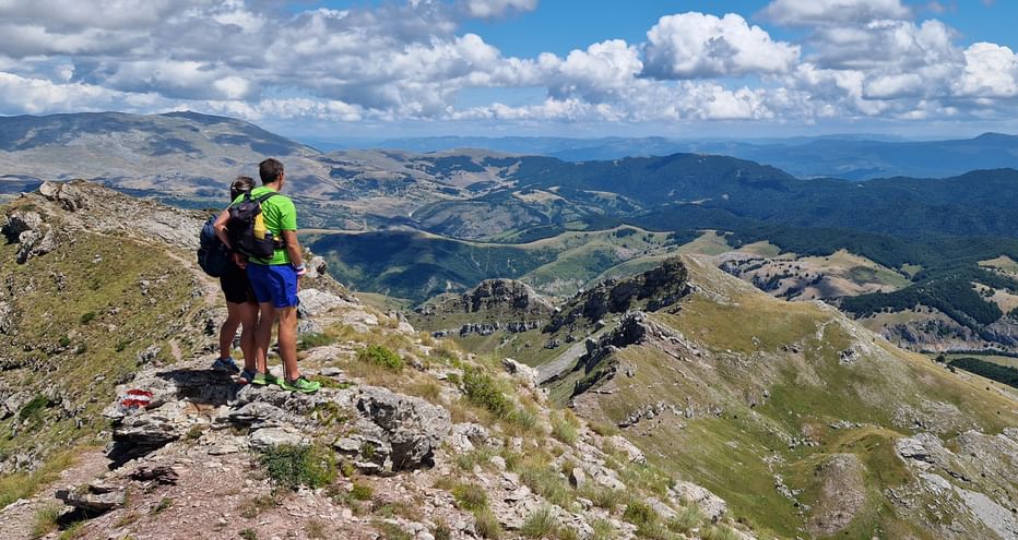 Two hikers with backpacks standing on rocky summit of Bjelasnica mountain in Bosnia, overlooking vast mountain landscape with valleys below.