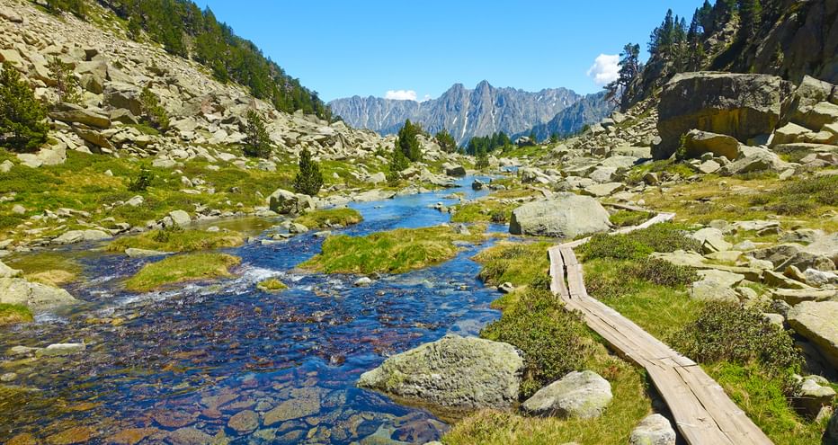Wanderpfad über Holzbrettern, entlang eines Baches inmitten von Wiesen und Felsen, mit Bergpanorama im Hintergrund