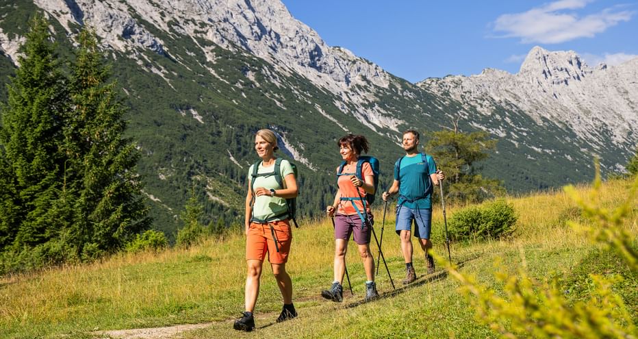Drei Wanderer mit Rucksäcken und Wanderstöcken auf einem Bergweg mit dramatischen Felsengipfeln und grünen Wäldern im Hintergrund.