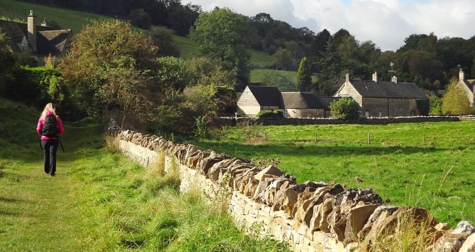 Hiker on a grassy path along a stone wall into a picturesque village of traditional stone houses