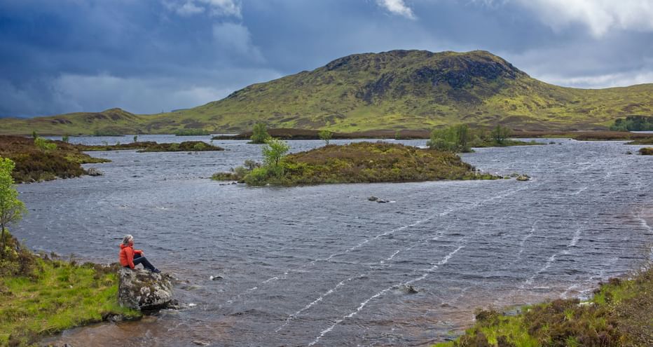Woman walker sitting on a large stone on the shore of a windswept loch of Rannoch Moor