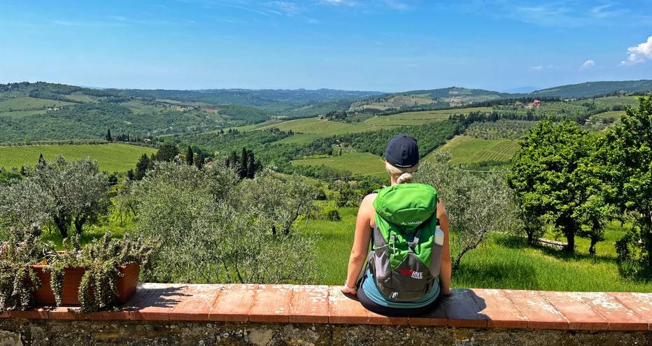 A hiker rests on a low wall in front of the church of San Leolino, with a view down the valley to the vineyards