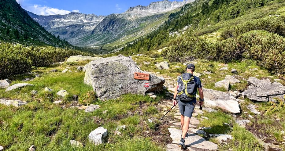 Wanderer mit Rucksack und Stöcken auf Bergweg im Krimmler Achental, Nationalpark Hohe Tauern. Wegmarkierung auf Felsen, Alpengipfel im Hintergrund.