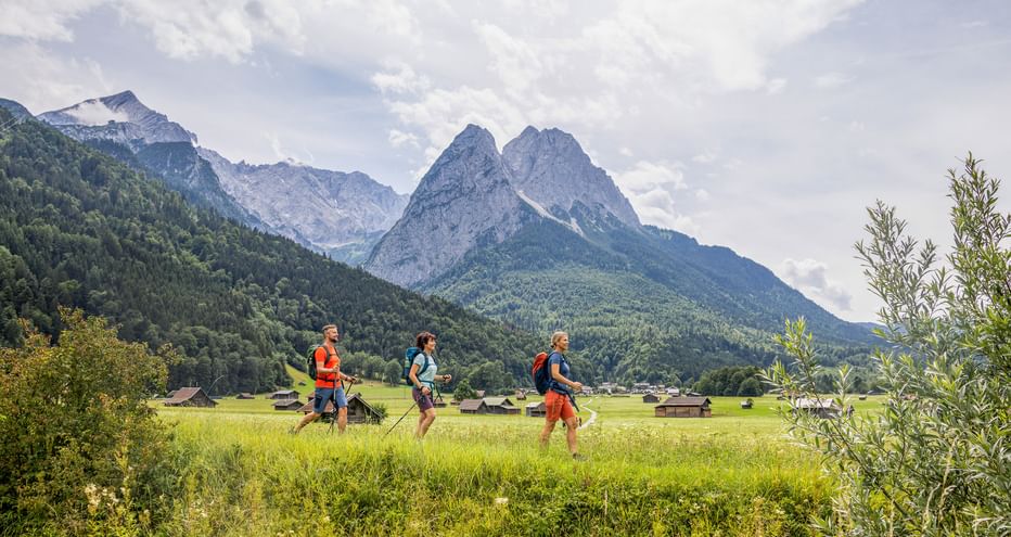 Three hikers with backpacks walking through green meadows near Garmisch with dramatic Alpine peaks in the background under cloudy sky.