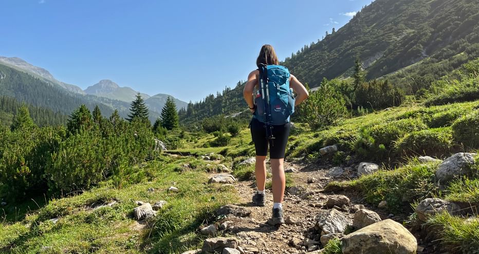 Wanderer mit blauem Rucksack auf felsigem Bergweg durch grünes Alpental mit bewaldeten Hängen und Berggipfeln unter blauem Himmel.