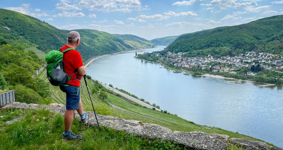 Wanderer mit Rucksack und Wanderstöcken auf Hang mit Blick auf Rheintal mit grünen Weinbergen und Dorf unter blauem Himmel.