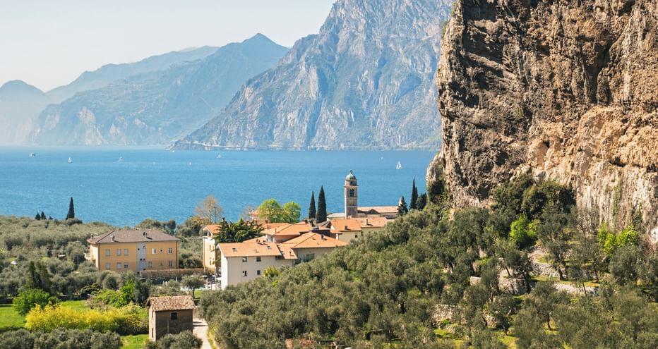 Torbole village with church tower nestled between dramatic cliffs and Lake Garda, surrounded by olive groves and cypress trees.