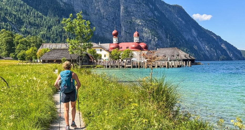 Wanderin mit Rucksack auf Uferweg am Königssee. St. Bartholomä Kirche mit roten Kuppeln über türkisfarbenem Wasser, Berge im Hintergrund.