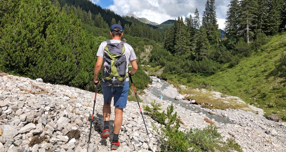 Wanderer am steinigen Bachbett mit Wald und Bergen im Hintergrund